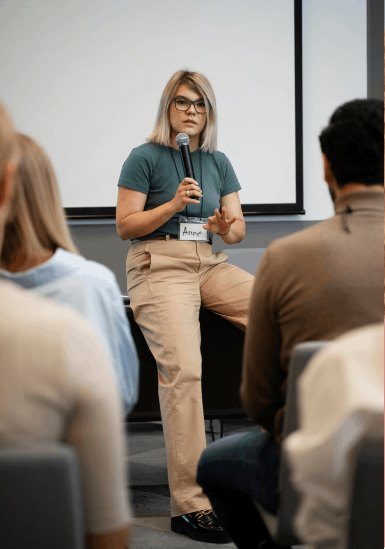 Une femme portant des lunettes et un badge nommé Anne parle avec un microphone devant un groupe de personnes lors d'une conférence ou d'un atelier.