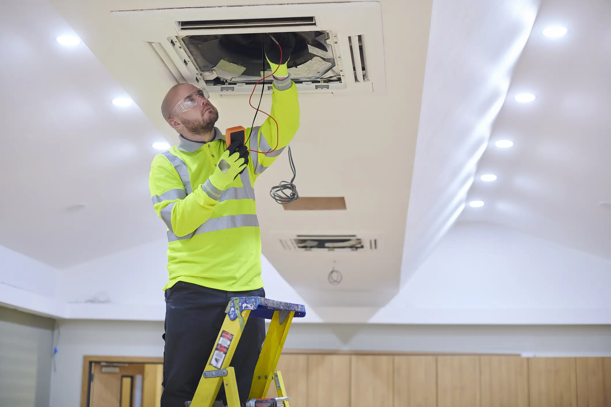 A technician wearing a high-visibility yellow-green shirt, safety glasses, and gloves is standing on a ladder. They are working on the wiring inside a ceiling-mounted cassette air conditioning unit, holding a device in one hand.