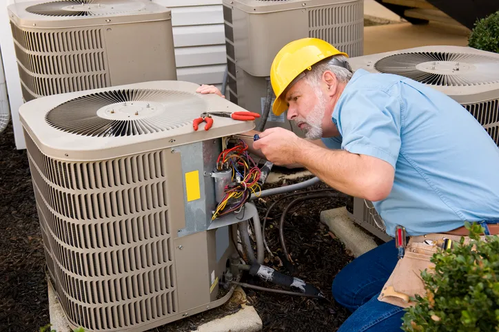 Technician in a hard hat repairing the electrical wiring inside an outdoor AC condenser unit.