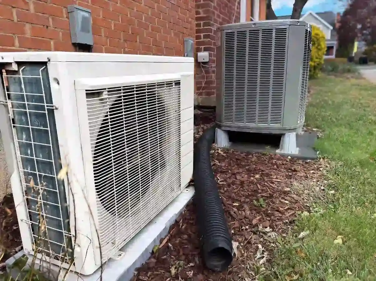 Two outdoor HVAC units beside a brick house: the left unit is older and white with visible dirt, while the right unit is newer and gray, elevated on concrete blocks. A black drainage pipe runs between them over mulch-covered ground.