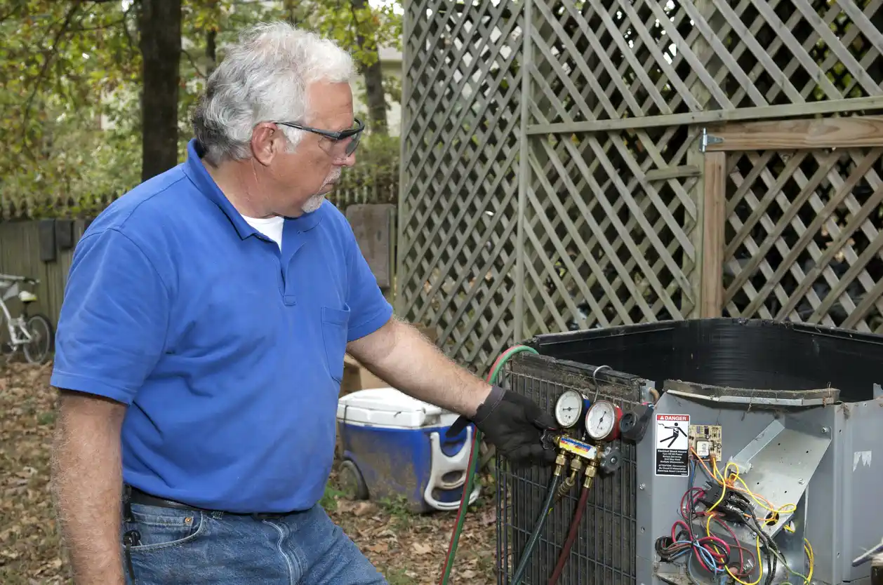 Technician in blue polo shirt, jeans, safety glasses, and one black glove inspecting manifold gauges connected to an open outdoor air conditioning unit. Internal wiring is exposed, with a warning label indicating electrical hazard. Residential setting includes a wooden fence and bicycle.