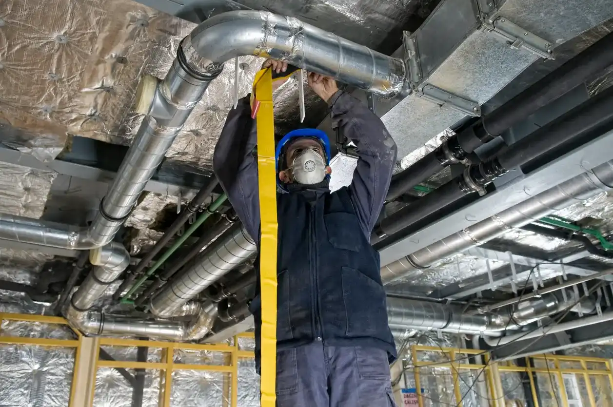 A construction worker wearing a hard hat and mask installs large metal ductwork on a ceiling.