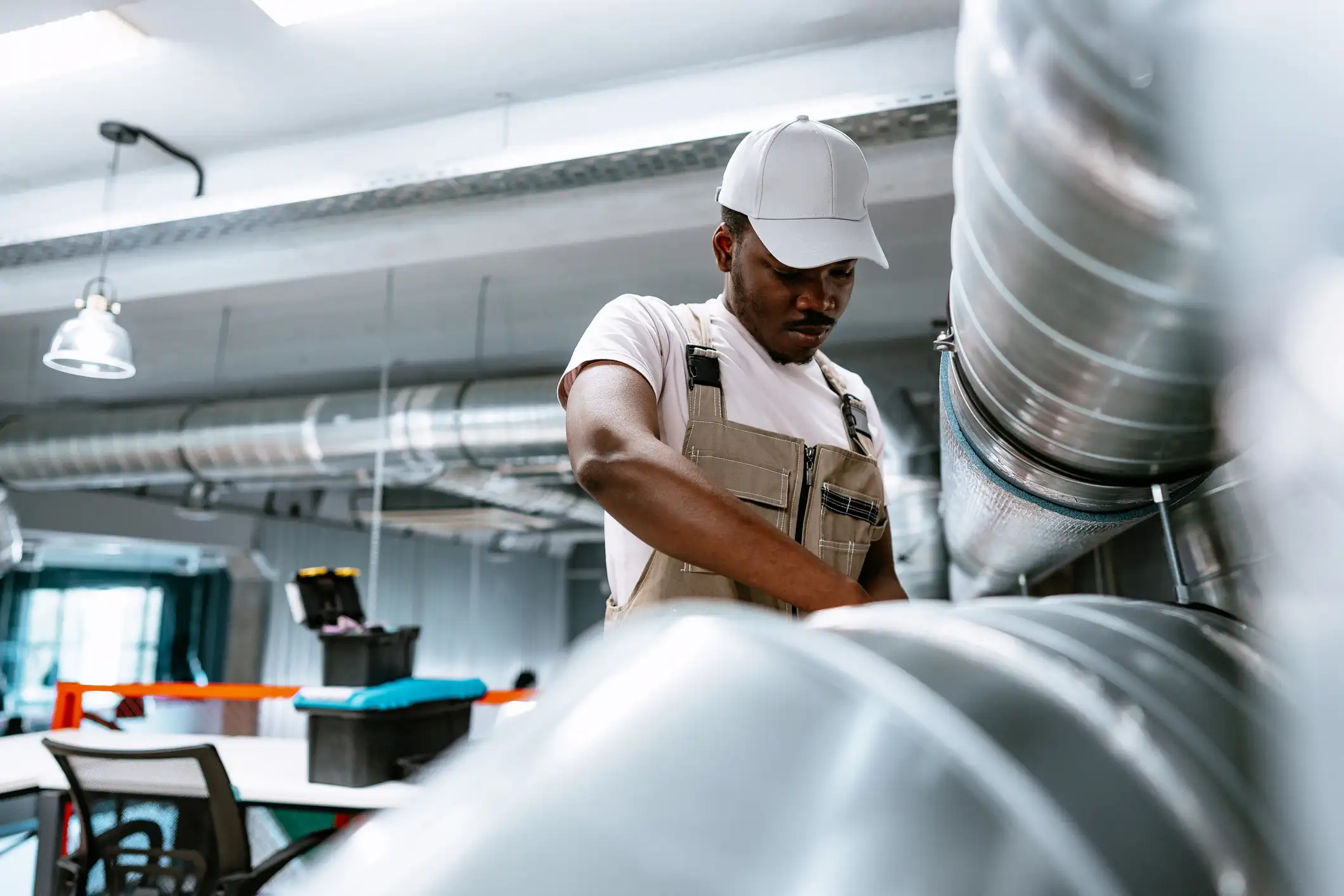 A young Black male technician in a white t-shirt, khaki overalls, and a white baseball cap is working on a large, silver, cylindrical air duct in a commercial or office space. He is framed by several parallel ducts in the foreground.
