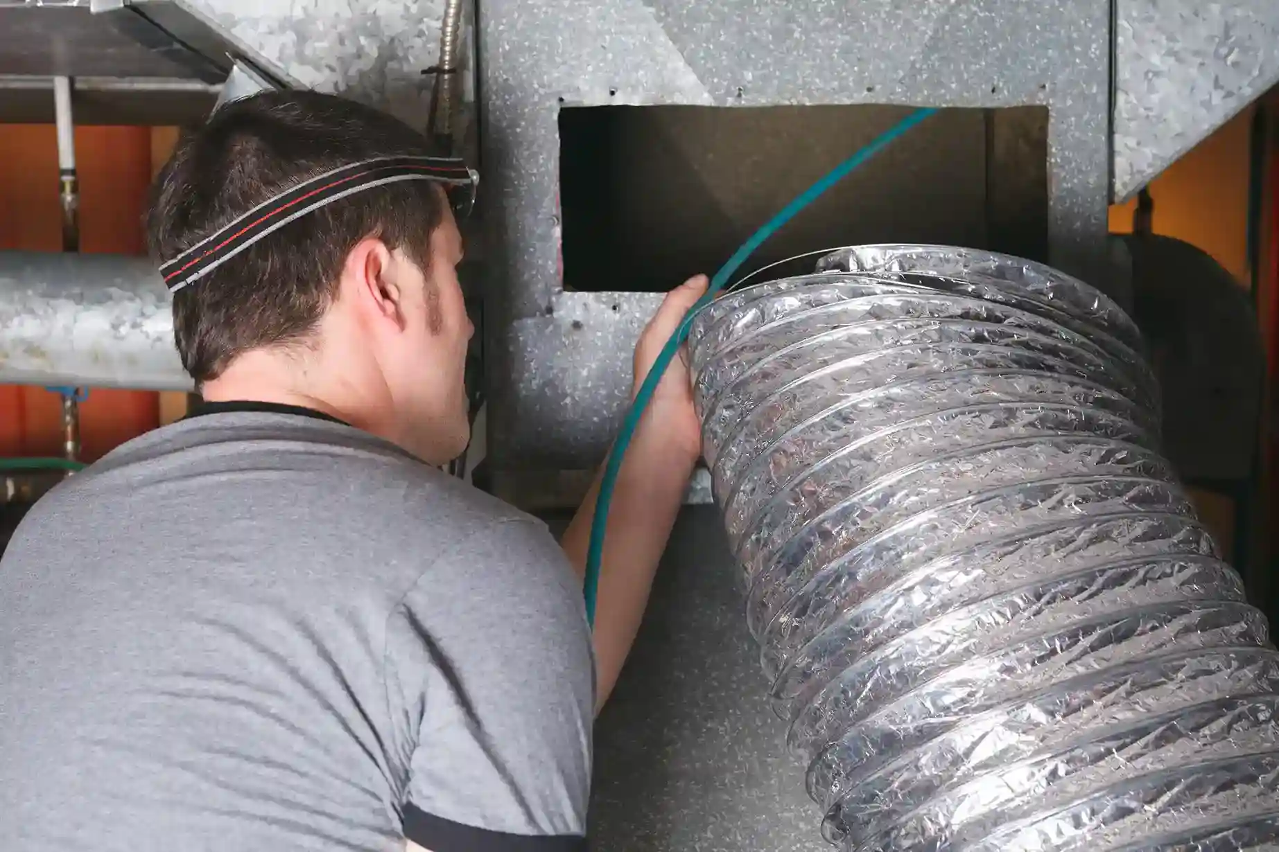 HVAC technician wearing a headlamp connecting flexible ductwork to a rigid metal air handler or plenum.