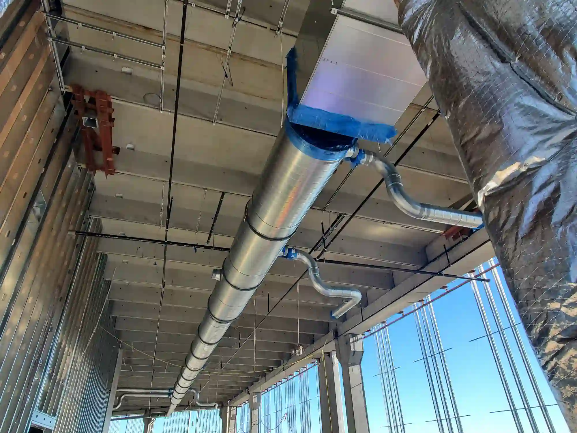 A view from below shows a long, central spiral galvanized metal air duct running across a concrete ceiling in a commercial or industrial space under construction. Smaller silver ducts branch off, and a large insulated duct is visible on the right.