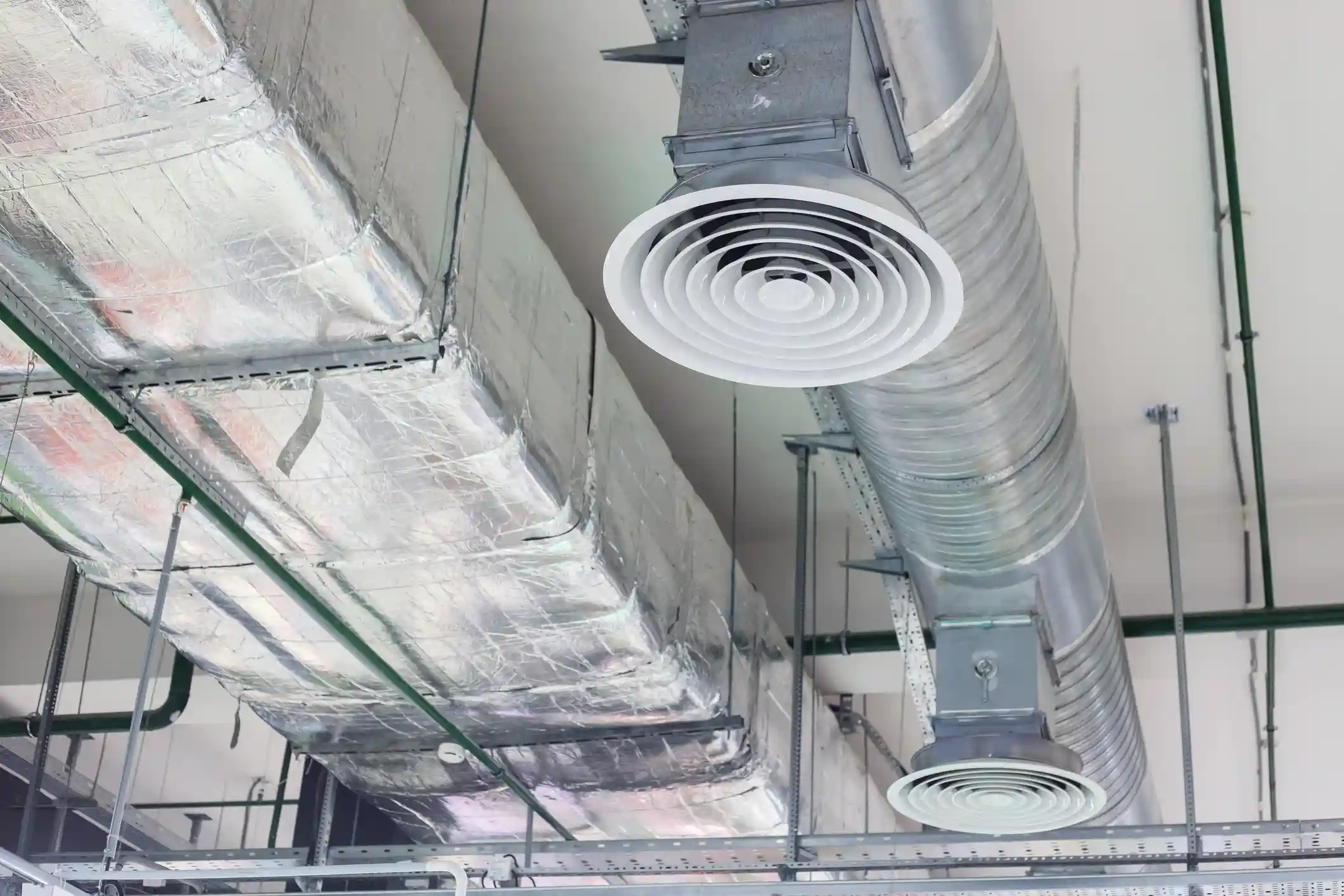 A low-angle view shows an insulated, rectangular aluminum air duct next to a cylindrical galvanized metal duct. Both ducts have large, round, white concentric circular air diffusers mounted beneath them, suggesting a commercial HVAC system.