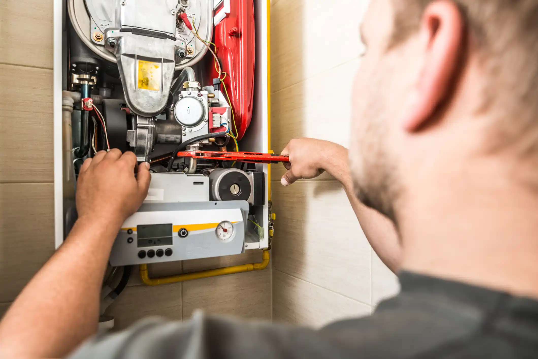 A technician with a beard and dark shirt adjusts the internal components of a wall-mounted red and white boiler unit. He uses a wrench on a part while his other hand reaches inside the open front panel, which exposes the heat exchanger and control mechanisms. The background is tiled.