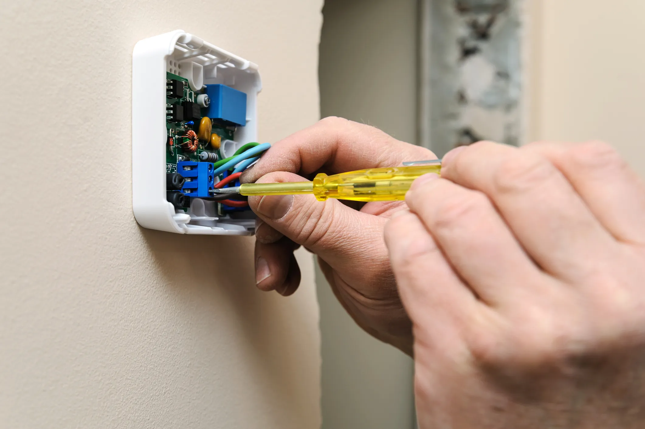 A person using a yellow screwdriver to tighten wiring on a small white wall-mounted thermostat circuit board