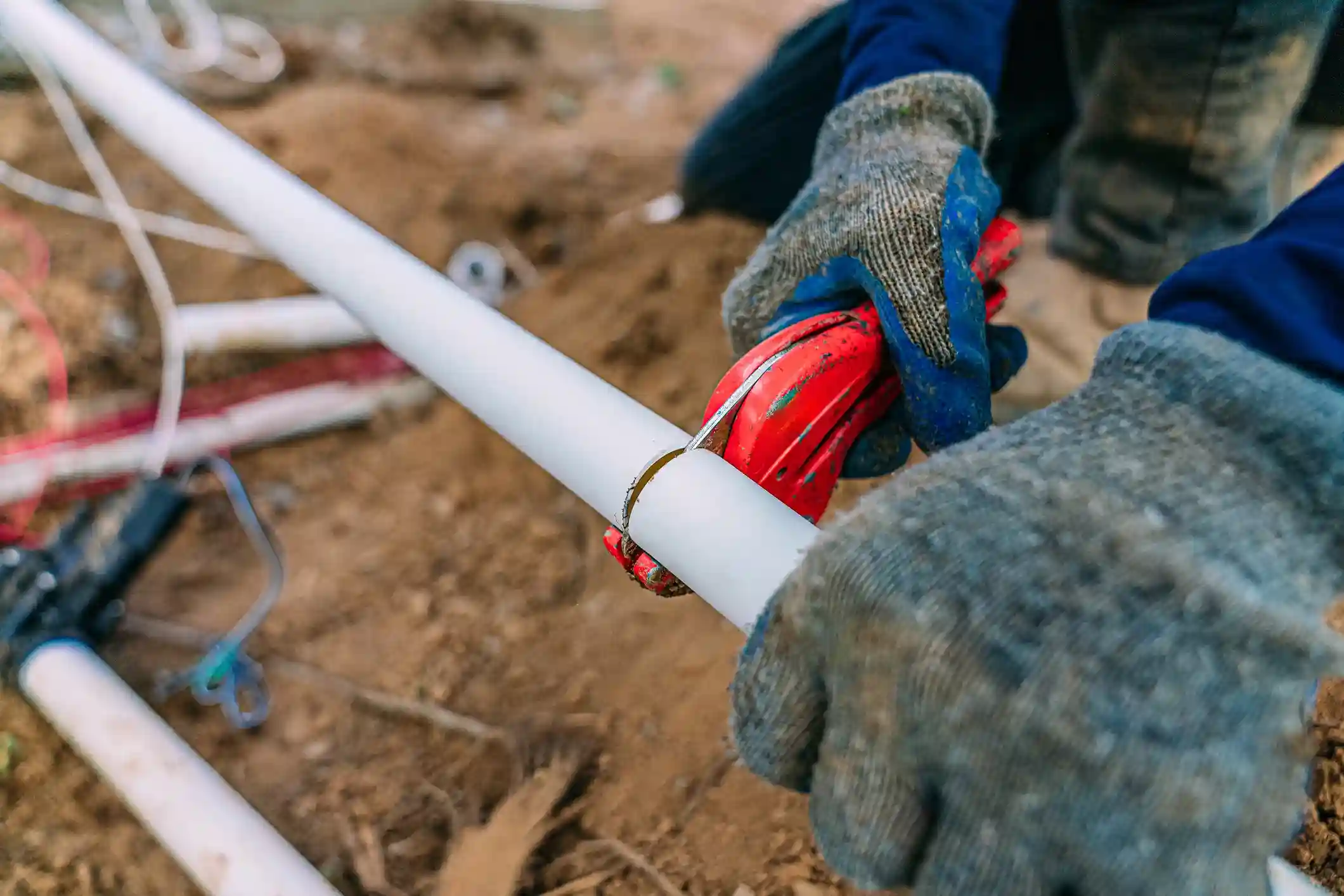 Hands in work gloves cutting a white PVC pipe outdoors for a plumbing repair.