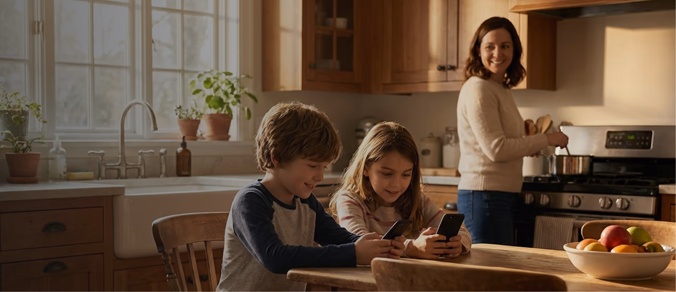 Two children sitting at a wooden kitchen table using smartphones while a woman cooks at the stove in the background.