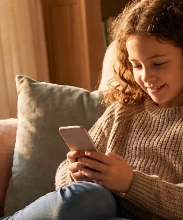 Young girl sitting on a couch, smiling while looking at a smartphone in her hands.