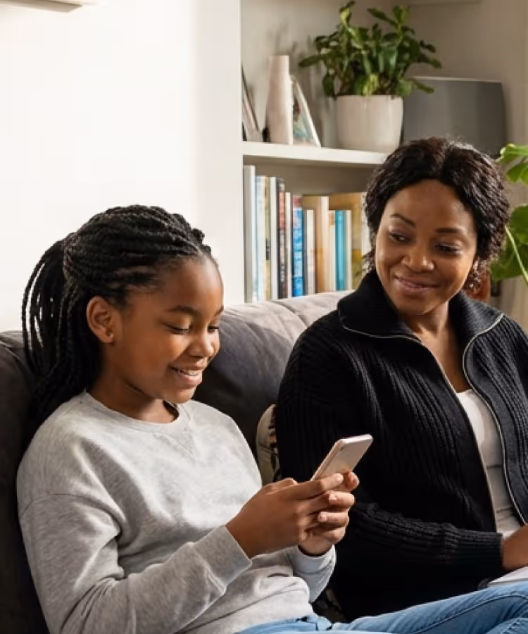Smiling mother and daughter sitting on a couch, with the daughter holding a smartphone.