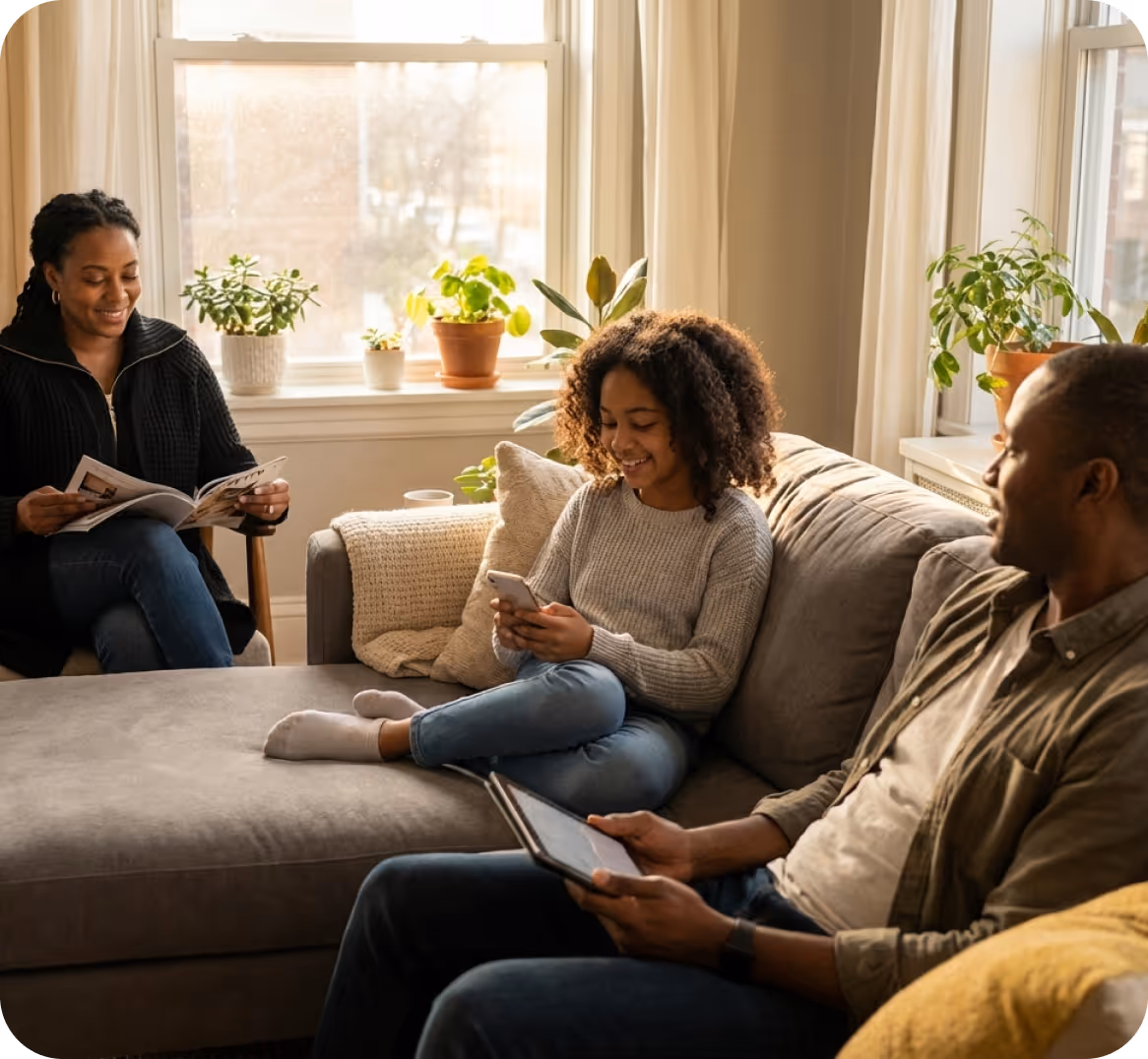 Family of three relaxing in a living room with natural light, the mother reading a magazine, the daughter using a smartphone, and the father holding a tablet.