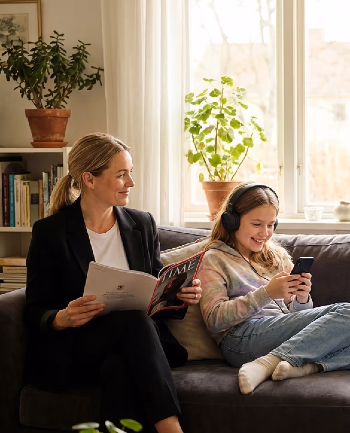 Woman reading a magazine and smiling at a girl wearing headphones who is using a smartphone on a couch.