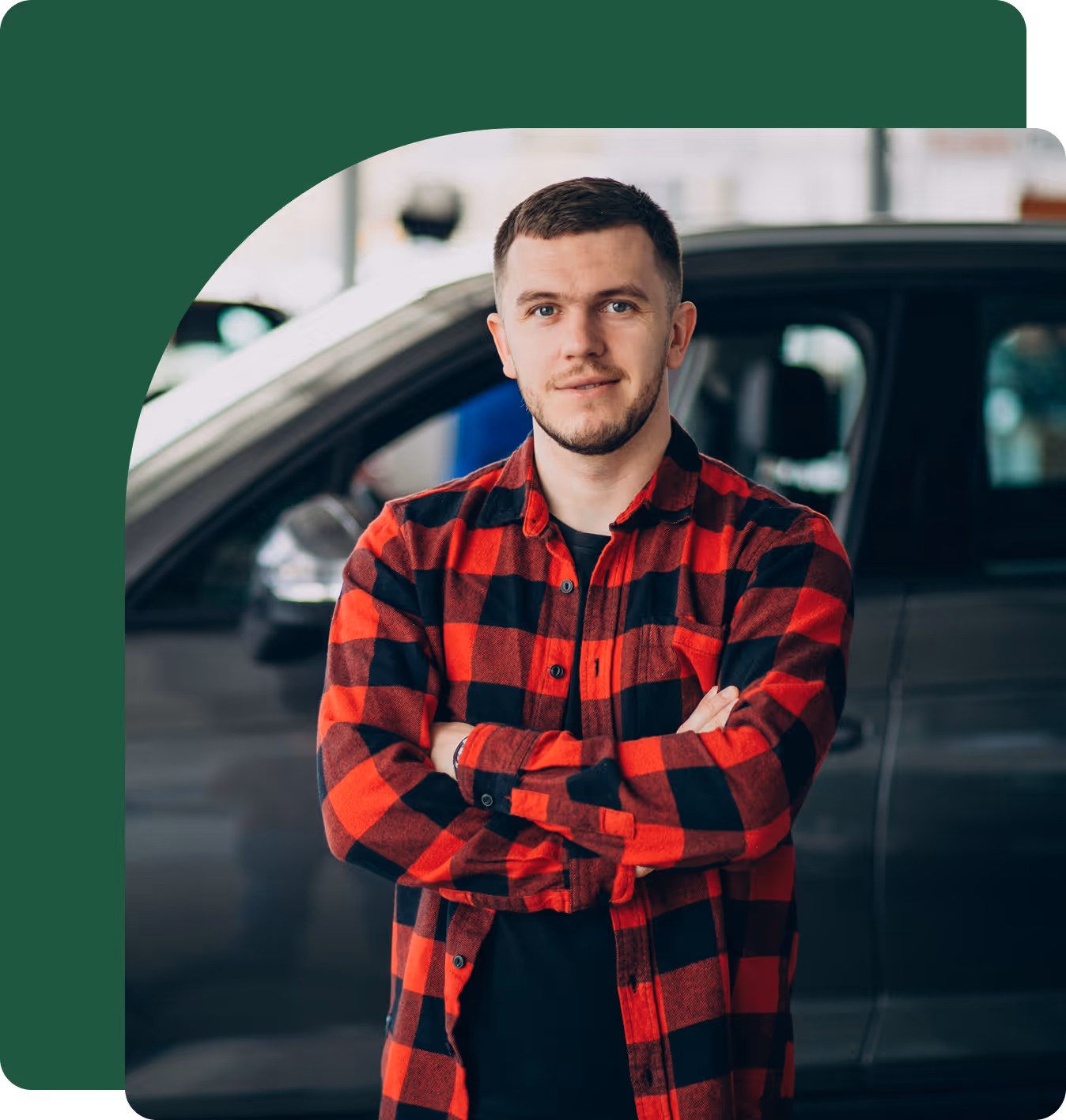 Man in red and black checkered shirt standing with arms crossed in front of a dark vehicle.