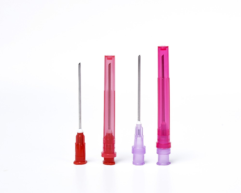 Close-up of four medical needles of different sizes, with red and pink plastic caps on a white surface.