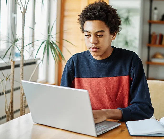 Young person with curly hair focused on a laptop while sitting at a wooden table with an open notebook.