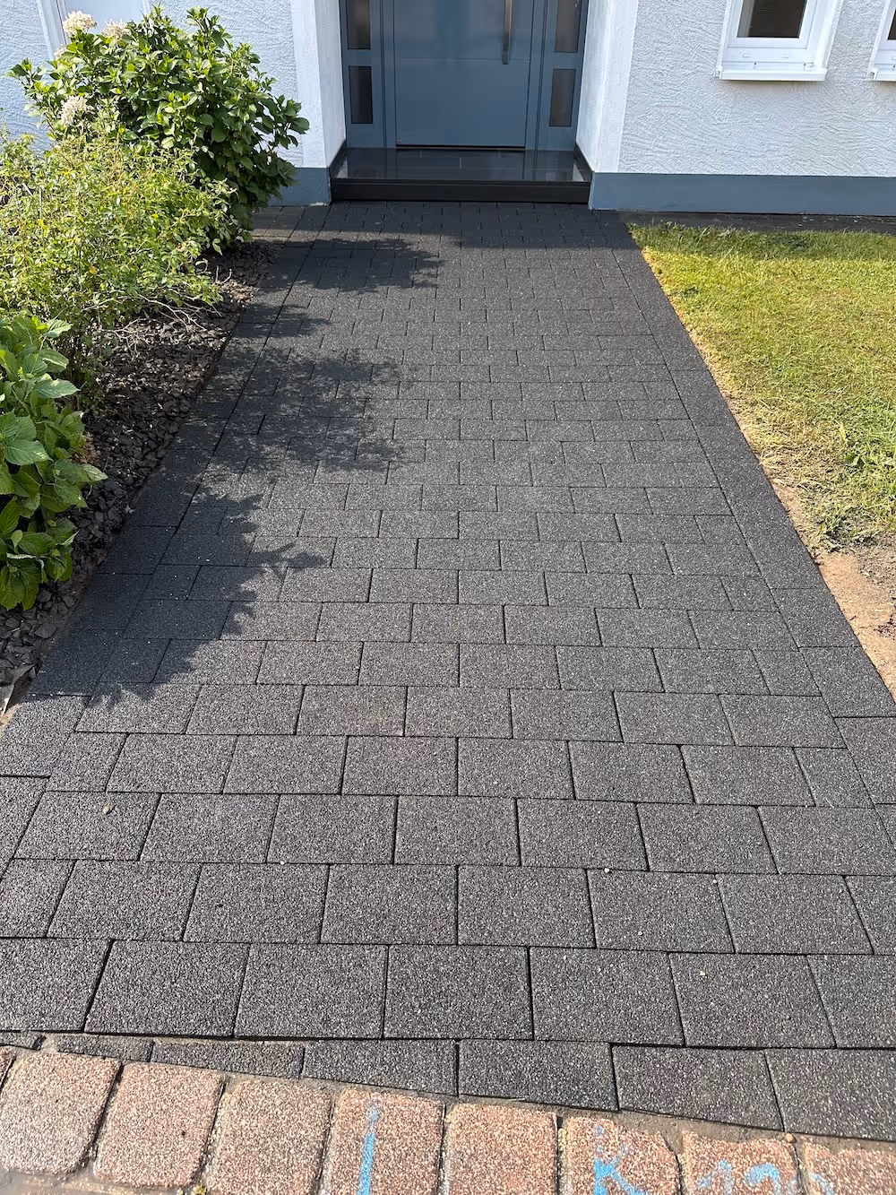 Sauber gefegter, dunkelgrauer gepflasterter Gehweg vor einem weißen Haus mit blauen Türen und Fenstern, links und rechts von Büschen und Rasen gesäumt.