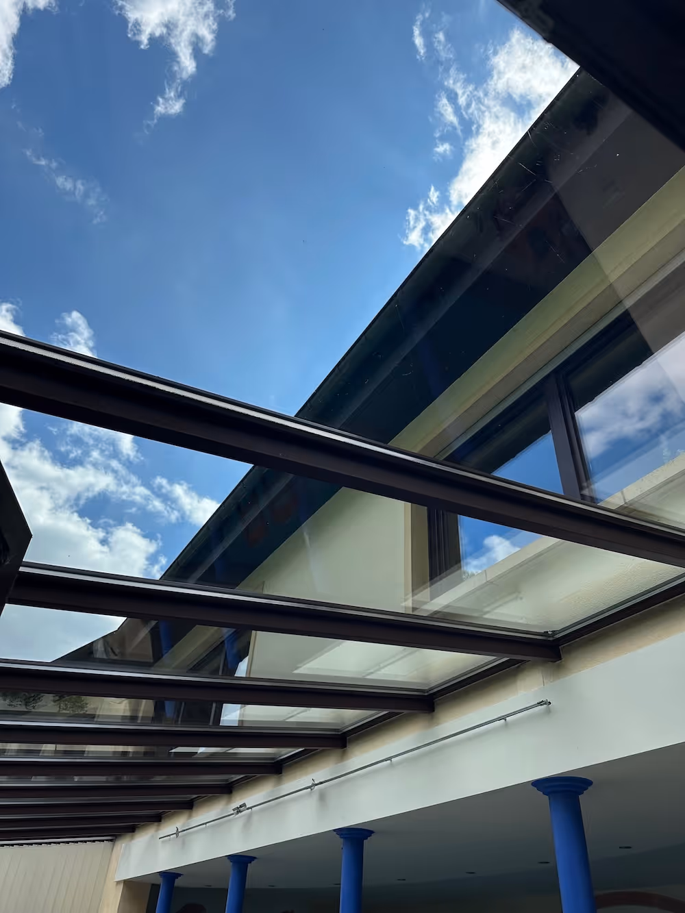 Glasdach mit schwarzen Metallstreben über einer Terrasse, blauer Himmel und Wolken im Hintergrund.