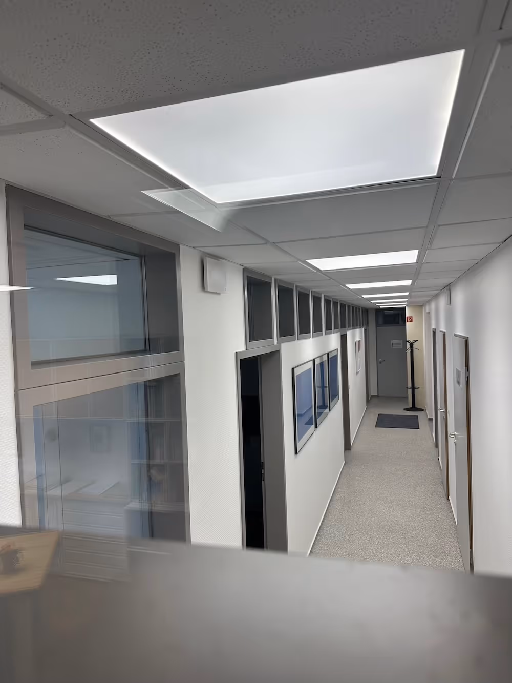 Moderner Flur in einem Bürogebäude mit weißen Wänden, mehreren geschlossenen und offenen Türen, Bildern an der Wand und Deckenbeleuchtung.