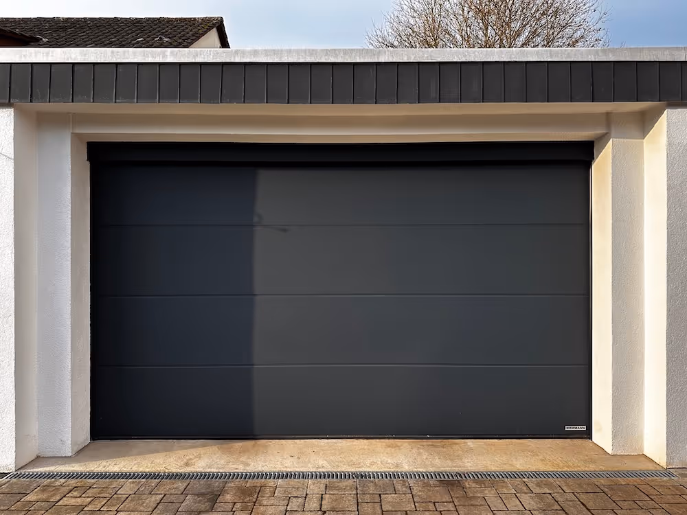 Modernes, geschlossenes schwarzes Garagentor in einem weißen Rahmen mit gepflastertem Zufahrtsbereich.