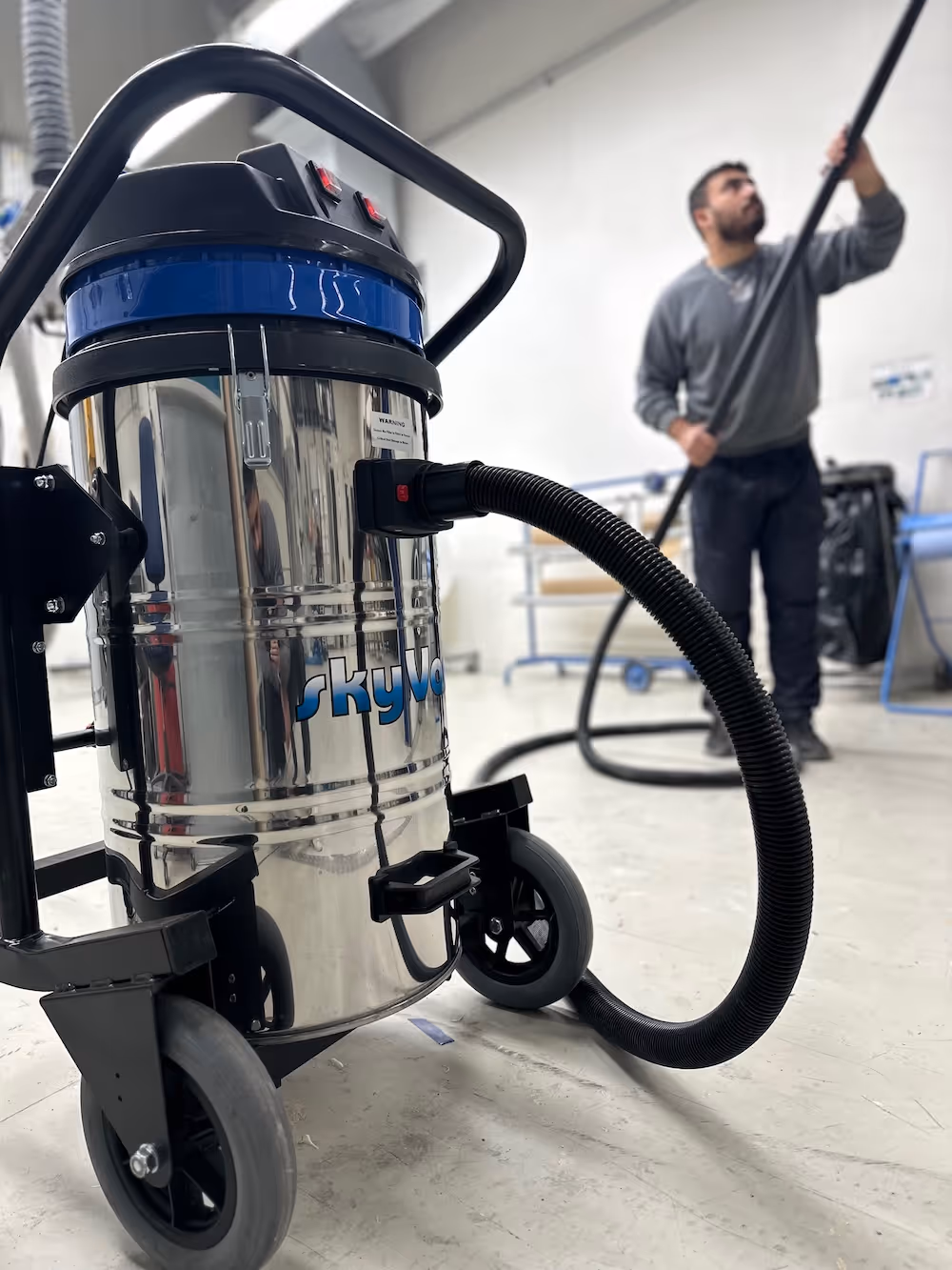 Industriesauger mit schwarzem Schlauch in einer Werkstatt, im Hintergrund reinigt ein Mann die Decke.