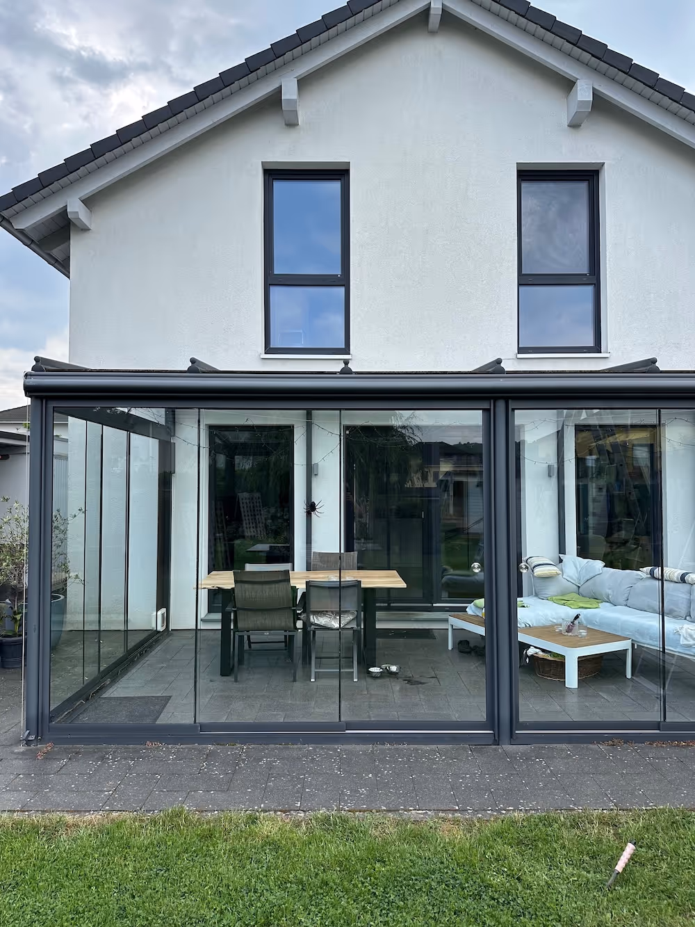 Moderner Wintergarten mit Glaswänden an einem weißen Haus, mit Esstisch, Stühlen und einer Sofaecke.