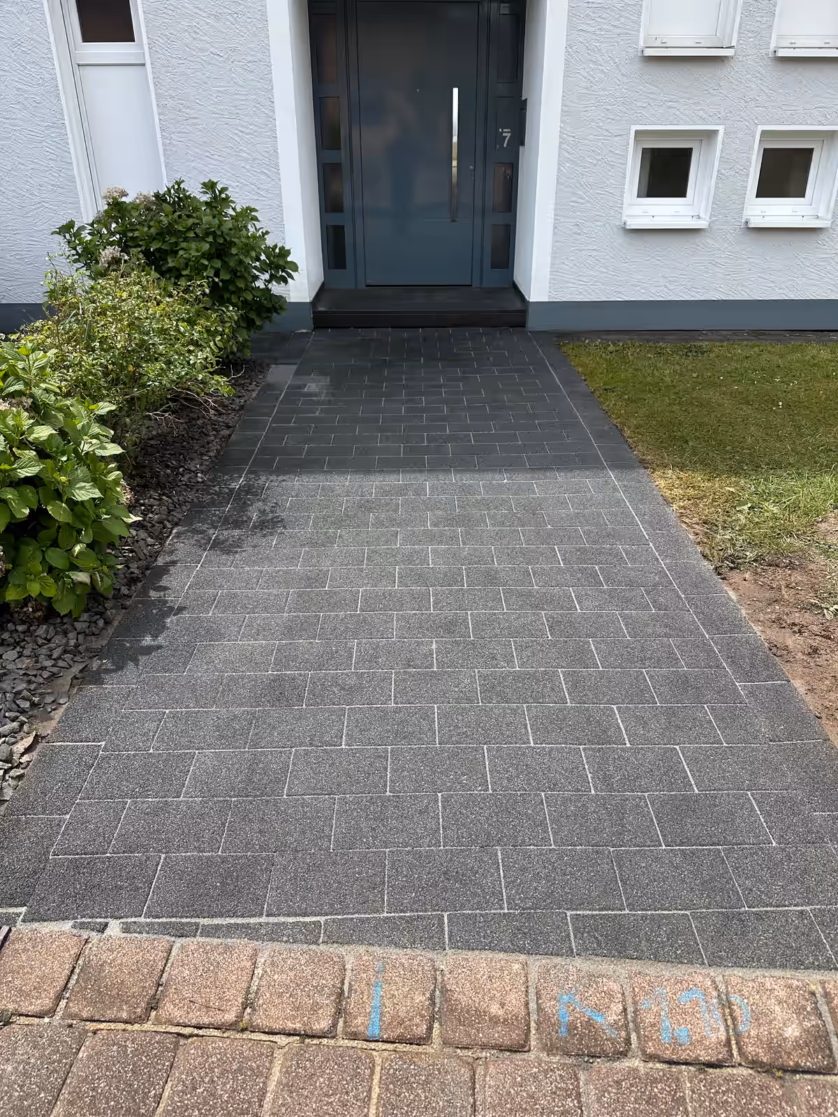 Eingangsbereich mit sauberen grauen Pflastersteinen, weißen Hauswänden, zwei kleinen Fenstern und grünen Pflanzen links.