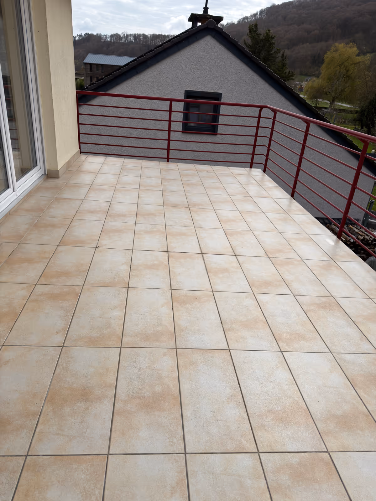 Hellbrauner gefliester Balkon mit rotem Metallgeländer und Blick auf ein graues Haus und bewaldeten Hügel im Hintergrund.