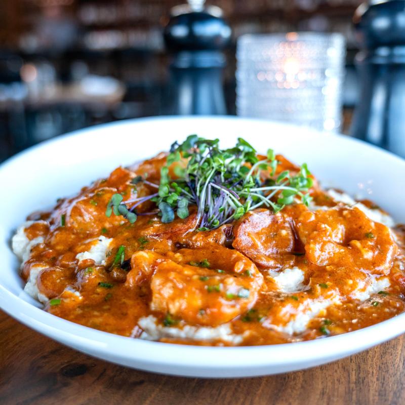 White bowl of shrimp in a creamy reddish-orange sauce served over grits, garnished with microgreens on a wooden table.