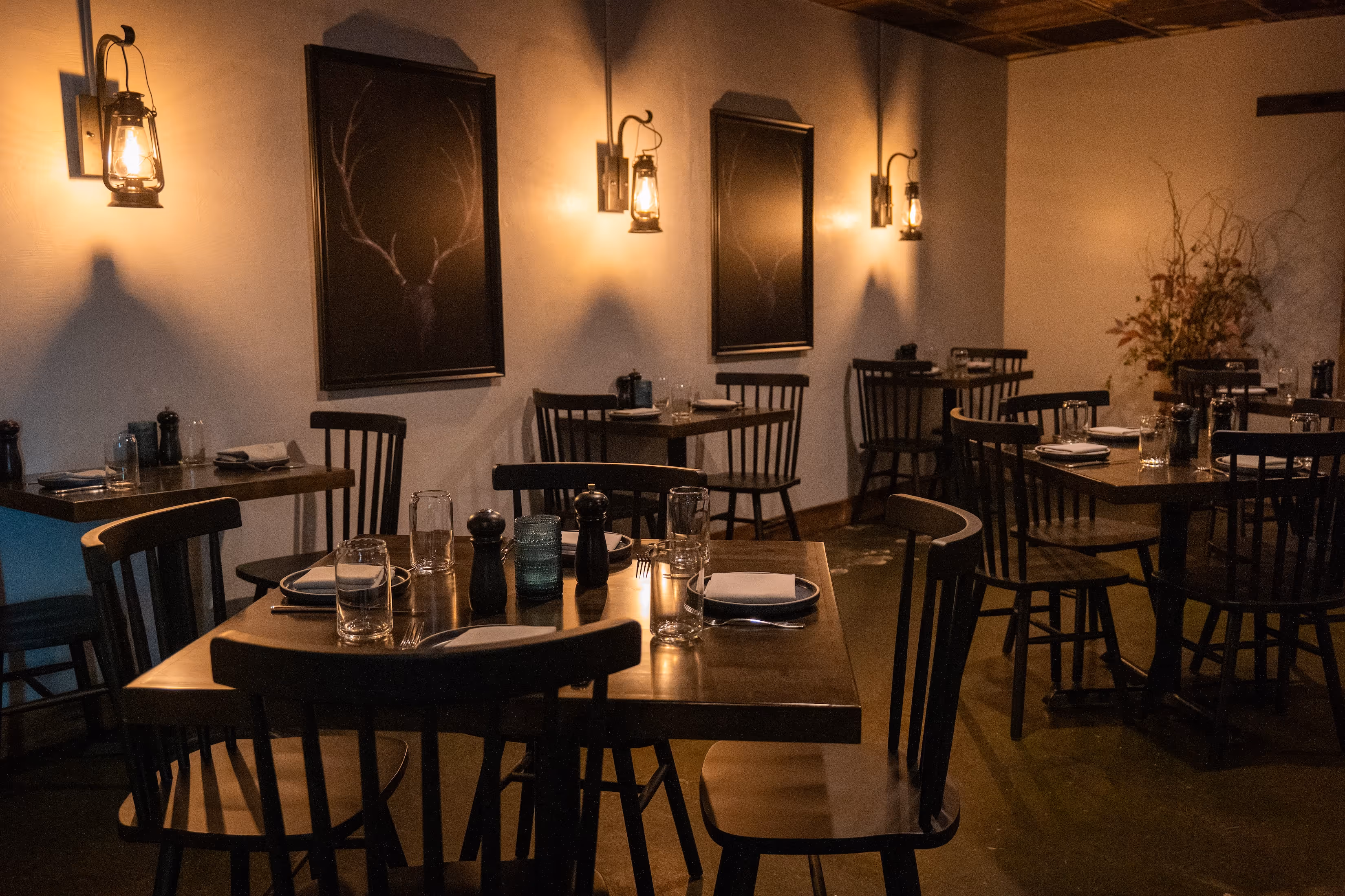 Dimly lit restaurant interior with wooden tables and chairs, lantern wall lights, and dark framed deer antler artwork.