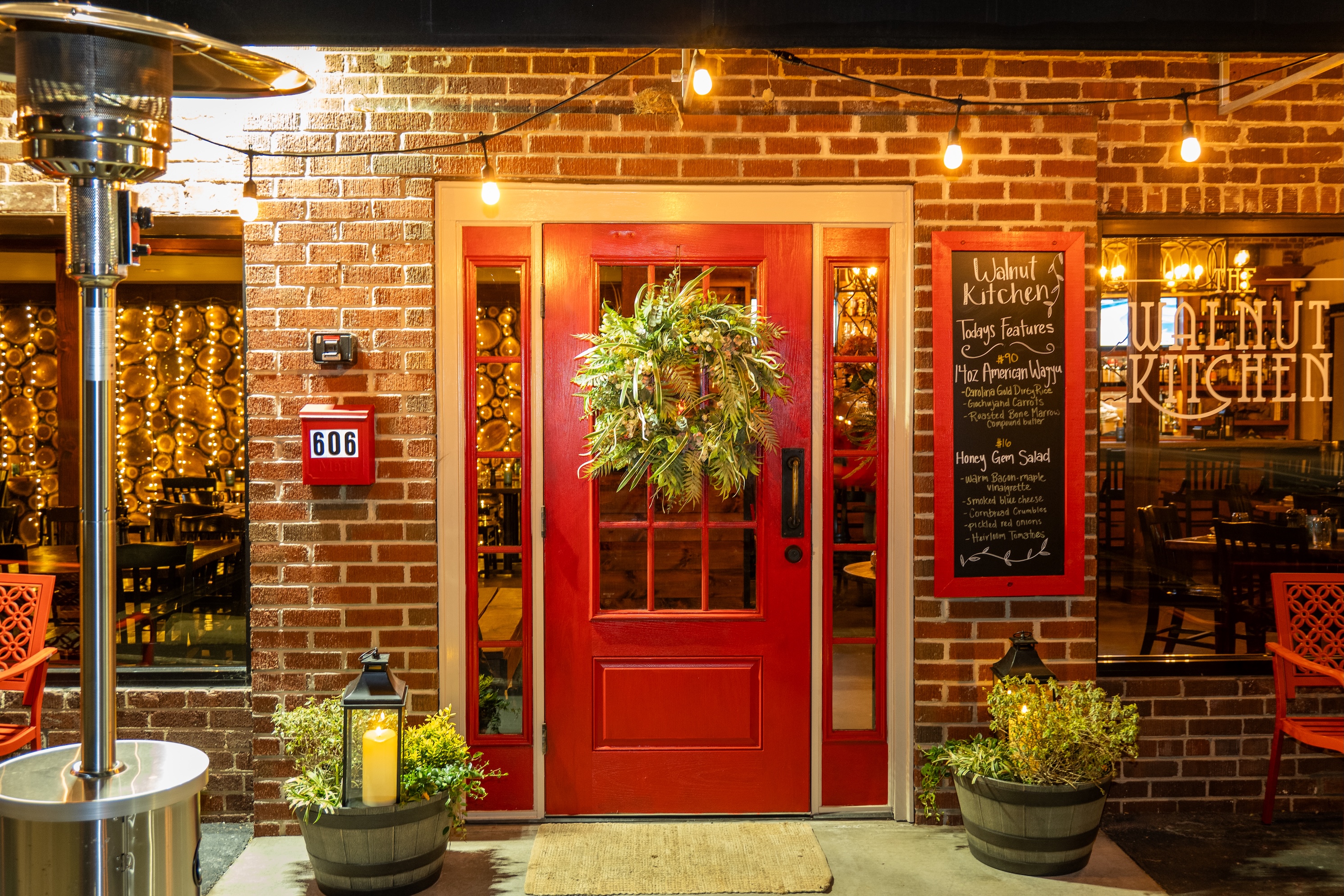 Cozy restaurant interior with wooden tables set with glasses, napkins, and salt and pepper shakers, a decorated Christmas tree, and large windows showing parked cars outside.