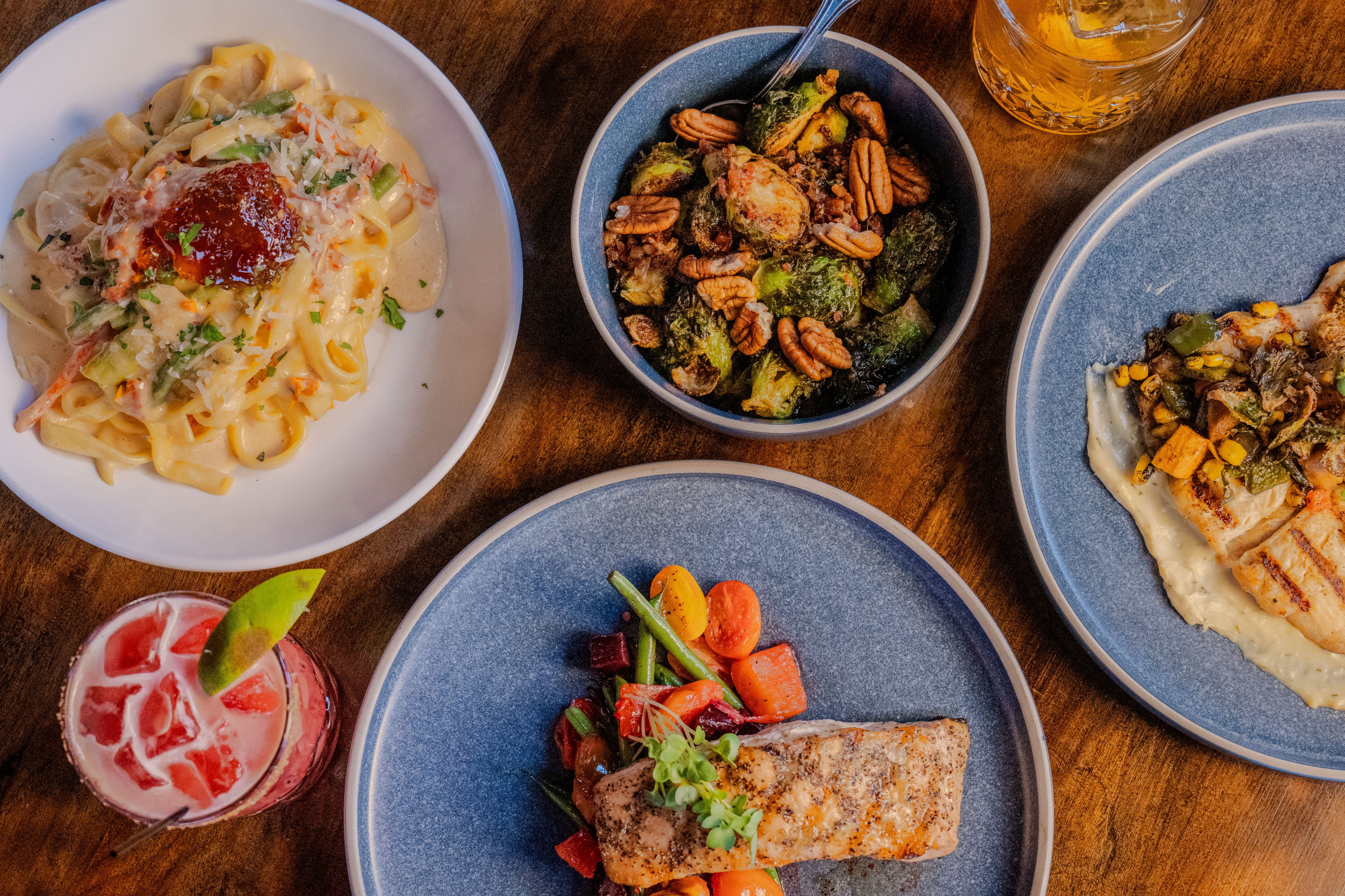 Wooden board with assorted cheeses, nuts, pickled vegetables, cured meats, biscuits, and sauces, with a glass of red wine and blurred lights in the background.
