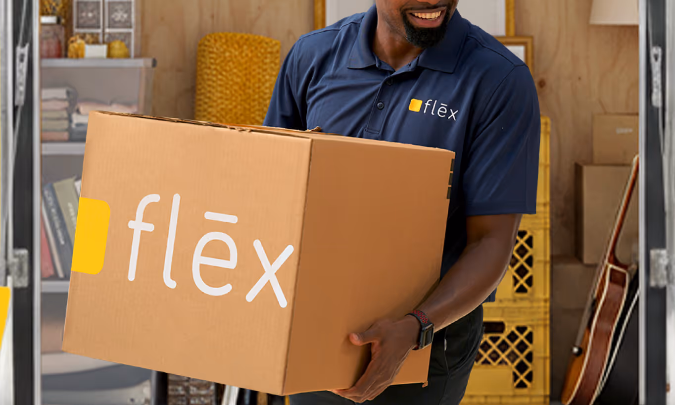 Smiling Black delivery worker in navy Flex uniform and cap carrying large cardboard box with yellow "Flex" logo on dark background.