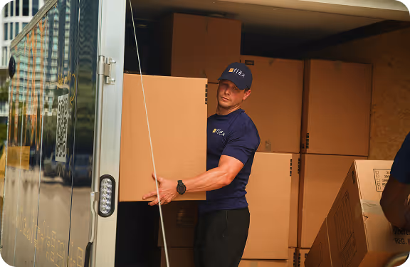 Muscular Flex team member in navy polo carrying a large cardboard box while exiting a blue & yellow branded trailer in an urban setting.