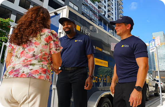 Flex team of two (two men in navy polos and caps) smiling and talking to one woman in floral top near a branded trailer in a city with high-rises.