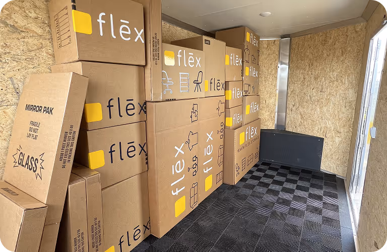Interior of a Flex trailer filled with stacked cardboard moving boxes labeled "Flex," "FRAGILE," "GLASS," and "MIRROR PAK" on plywood walls.