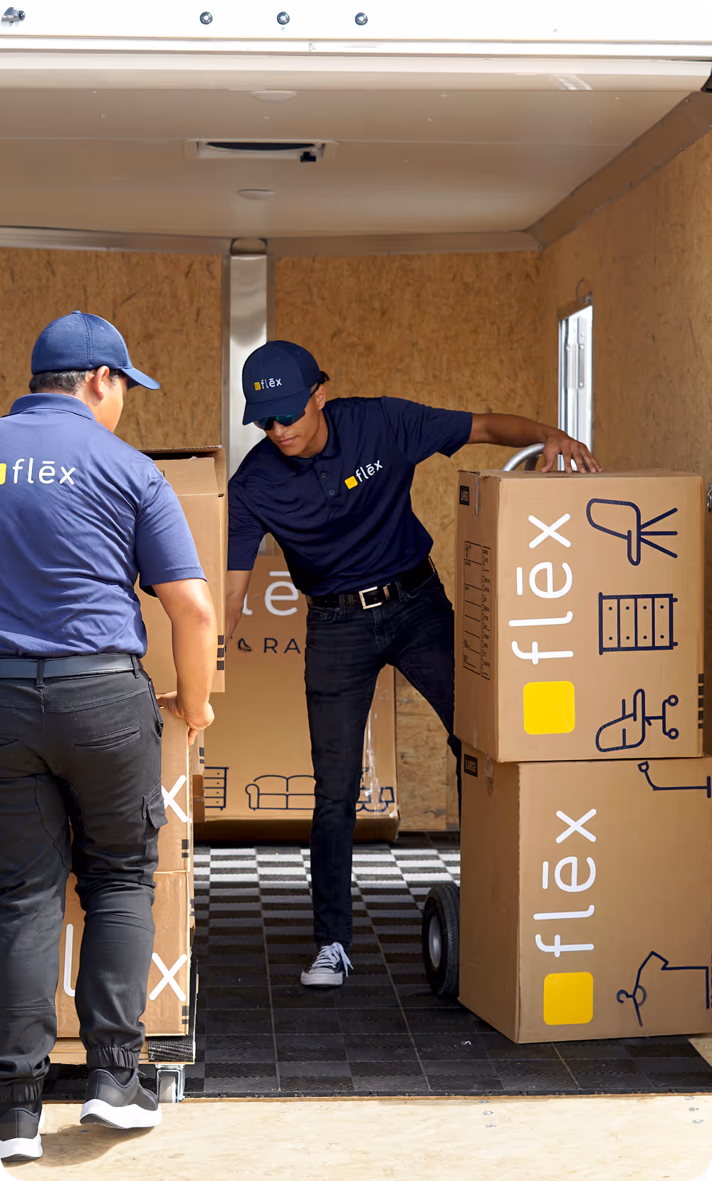 Two Flex workers in navy uniforms and caps carefully loading large labeled cardboard boxes inside a plywood-lined trailer interior.