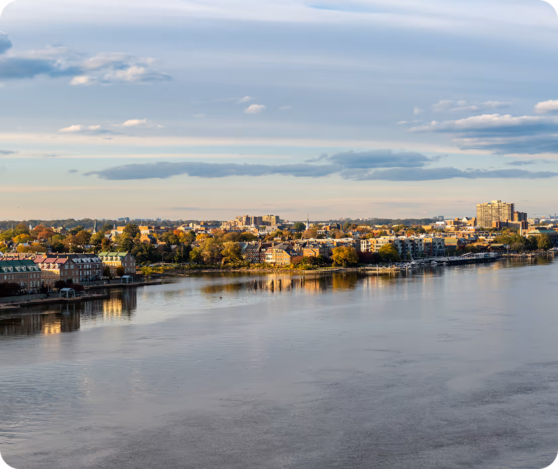 Aerial view of Richmond, Virginia skyline across the James River at dusk: mid-rise buildings and historic brick structures reflected in calm water, fall foliage in orange and green along the banks, under a soft blue-gray cloudy sky with golden light.