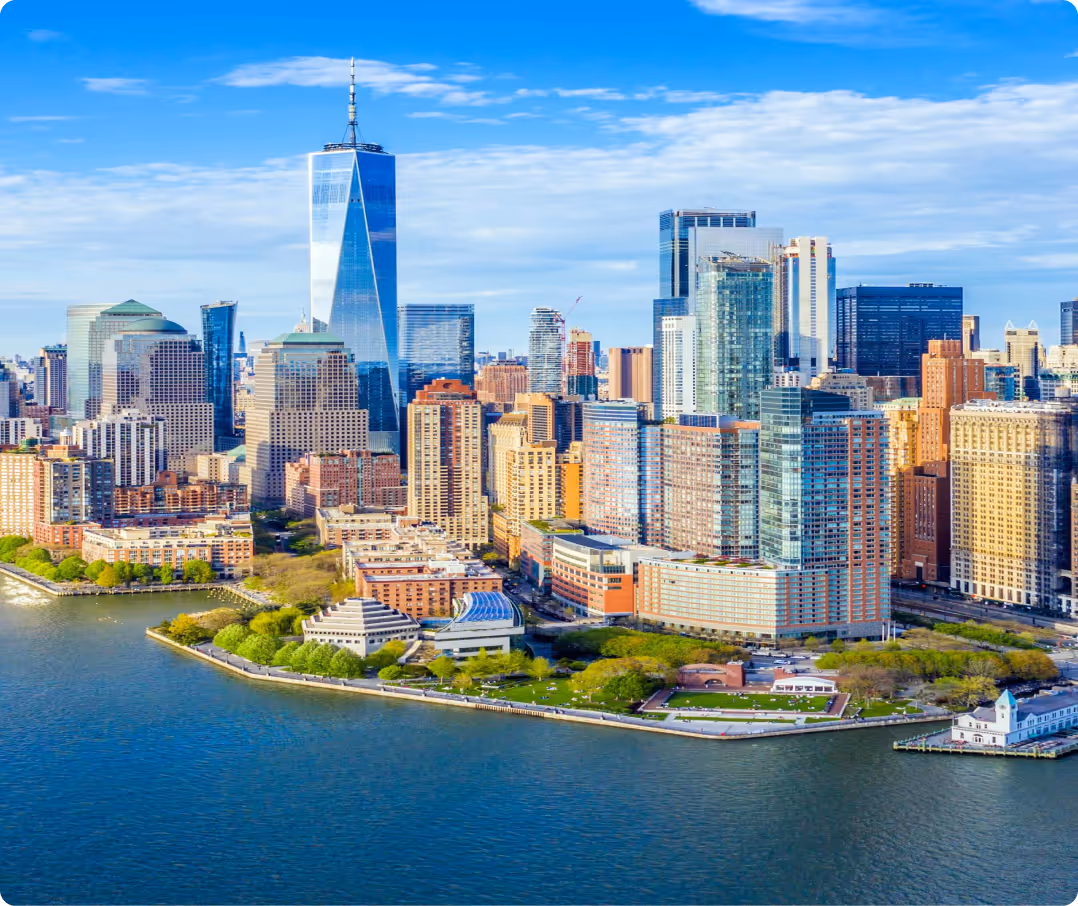 Aerial view of Lower Manhattan, New York City: One World Trade Center (Freedom Tower) rises prominently among dense skyscrapers, with the Hudson River, Battery Park greenery, and Brooklyn in the distance under a clear blue sky.