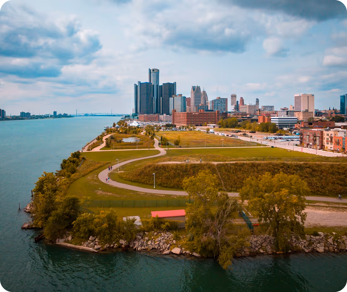 Aerial view of Detroit, Michigan skyline across the Detroit River: modern skyscrapers including the Renaissance Center towers rise behind a green park peninsula with winding paths, people walking, and rocky shoreline under a partly cloudy blue sky.