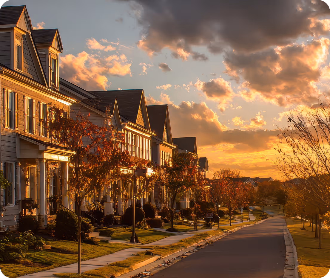 Golden hour sunset over a suburban Maryland neighborhood: row of two-story townhomes with gabled roofs, fall-colored trees lining the street, warm orange clouds in the sky, and quiet residential road in front.
