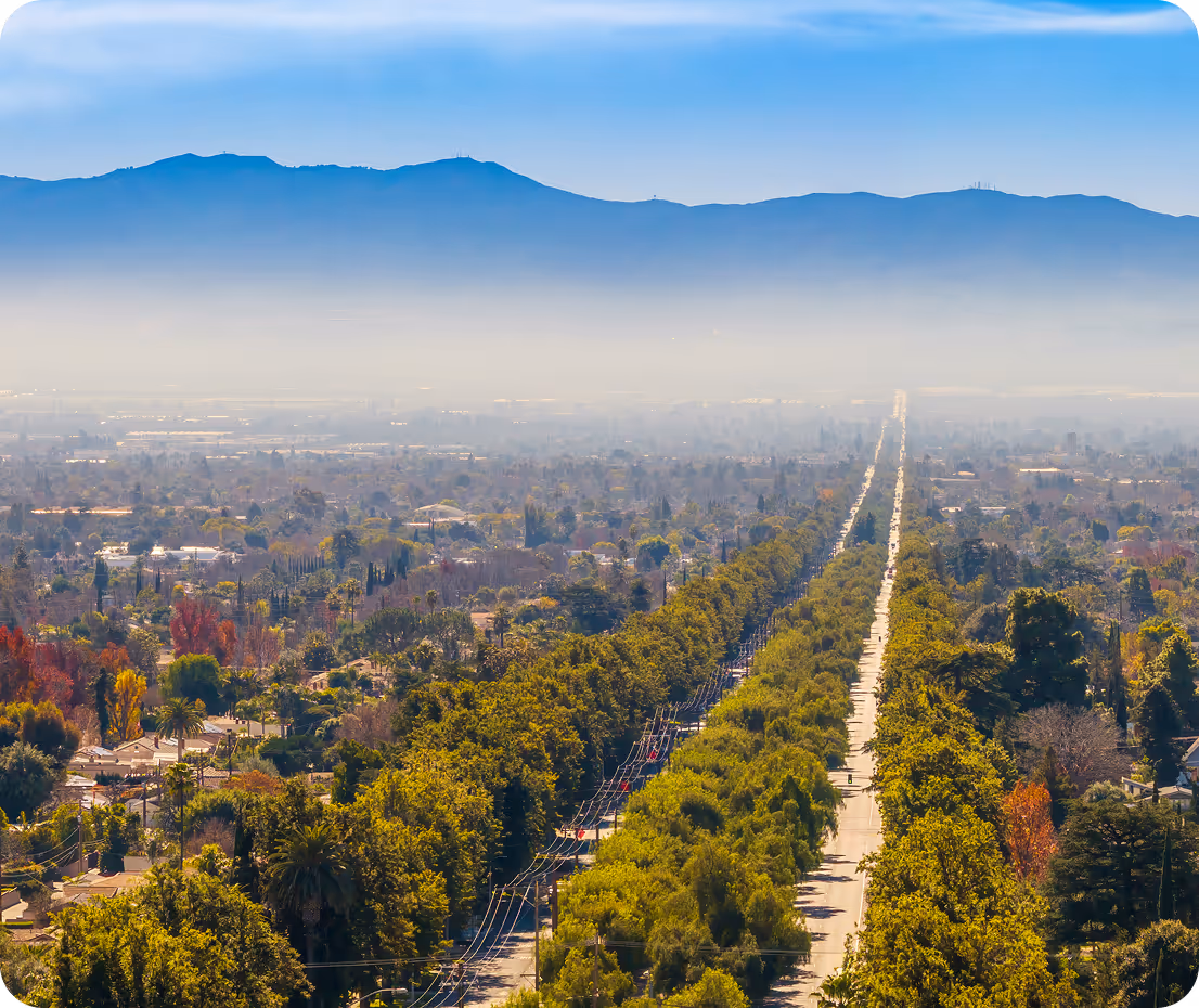 Aerial view of Los Angeles, California: a long, straight urban street lined with green trees stretches into the hazy distance toward the blue San Gabriel Mountains under a clear sky with light morning fog.
