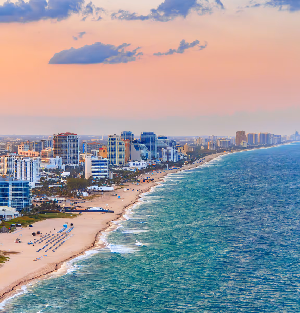 Aerial view of Fort Lauderdale beach with sandy shore, rows of beach umbrellas, ocean waves, and a skyline of high-rise buildings at sunset.
