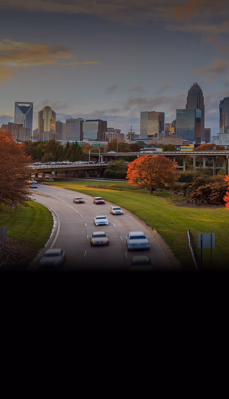 View of Charlotte NC skyline at sunset with highway and cars in foreground and autumn trees with colorful foliage.