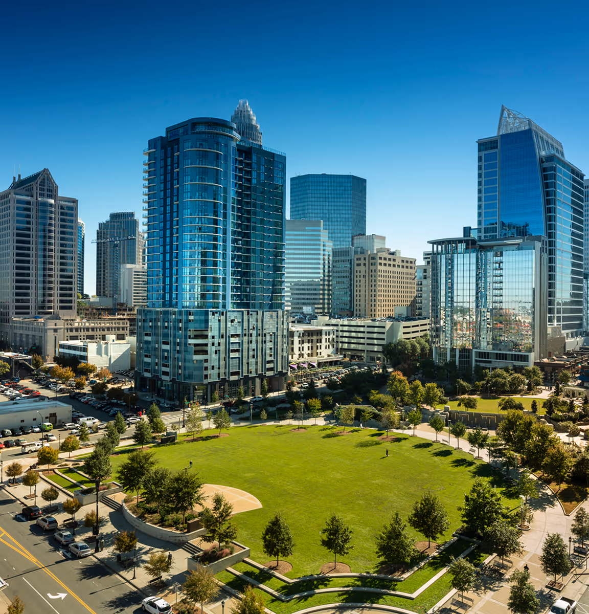 Aerial view of a city park with green lawn and trees surrounded by modern glass office buildings under a clear blue sky.