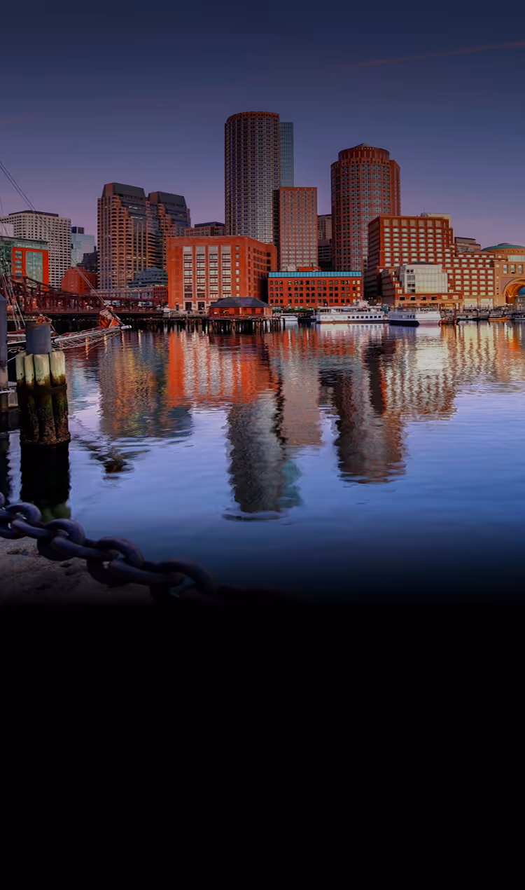 Boston skyline with tall buildings reflecting on calm waterfront at dusk.