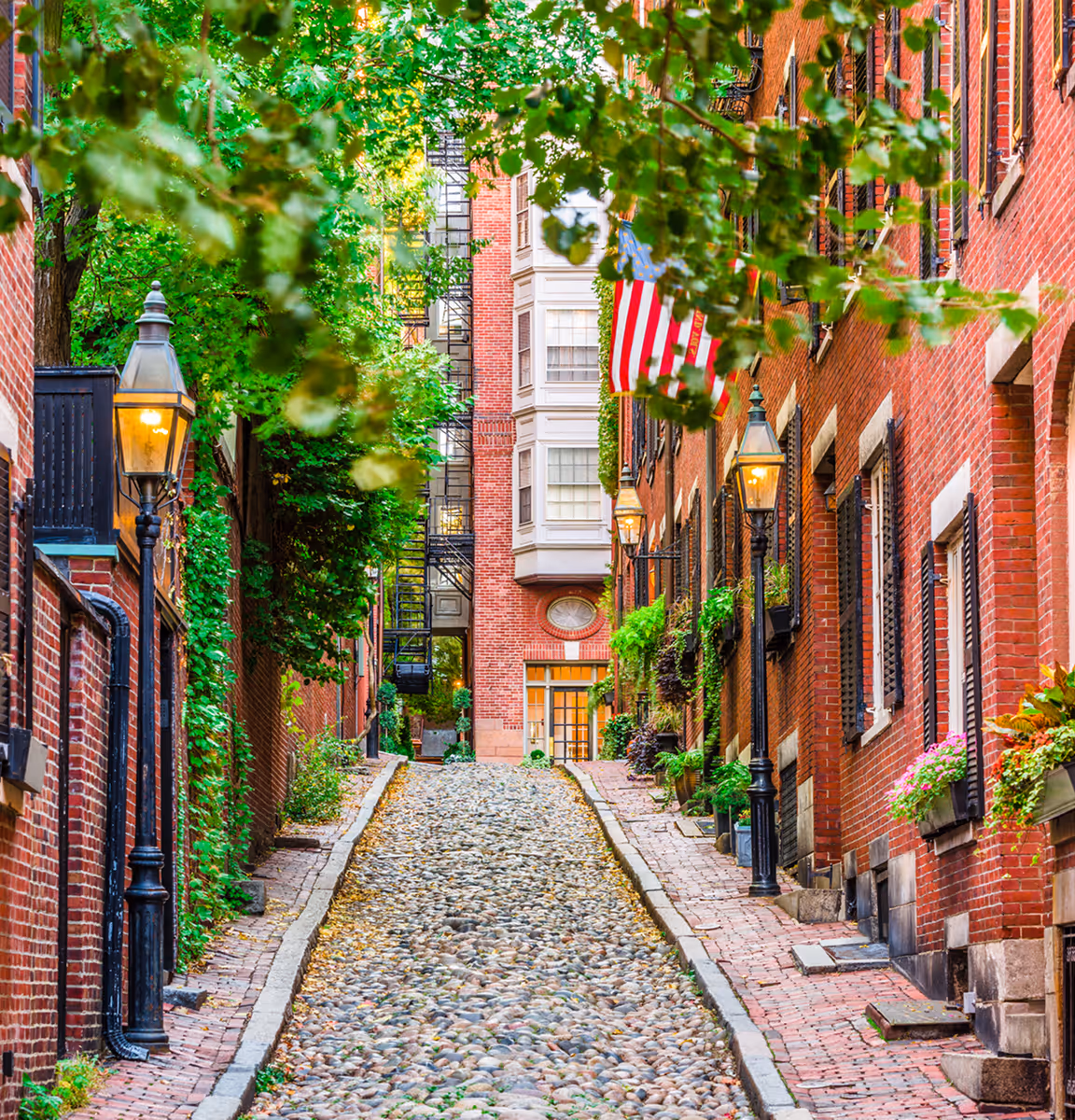 Narrow cobblestone street lined with red-brick buildings, green plants, and traditional street lamps with an American flag hanging on the right.