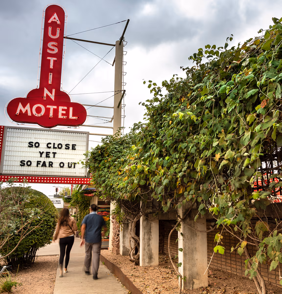 Two people walk on a sidewalk under a red Austin Motel neon sign with the message 'So close yet so far out', surrounded by green foliage.