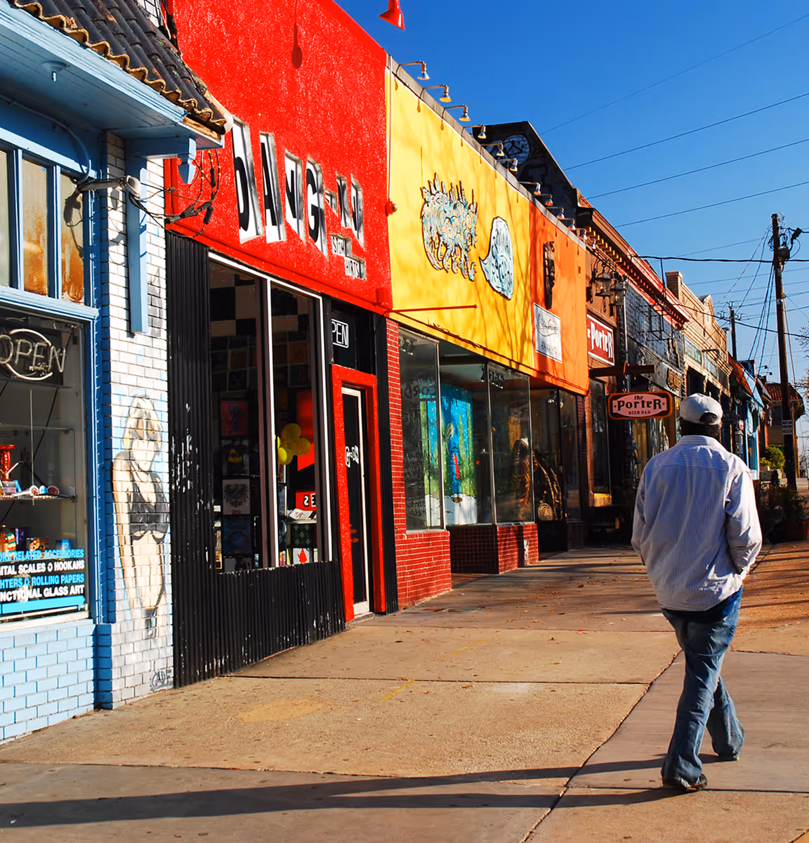 Man walking on sidewalk past colorful storefronts including a red building with large white letters and a yellow building with artwork under a clear blue sky.